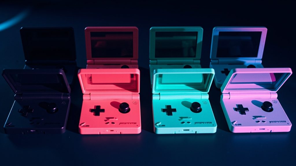 Illustration: Four clamshell-style handheld gaming consoles displayed side by side in black, pale pink, mint green, and silver, each featuring an upper screen, a directional pad, action buttons, and a small speaker grille.